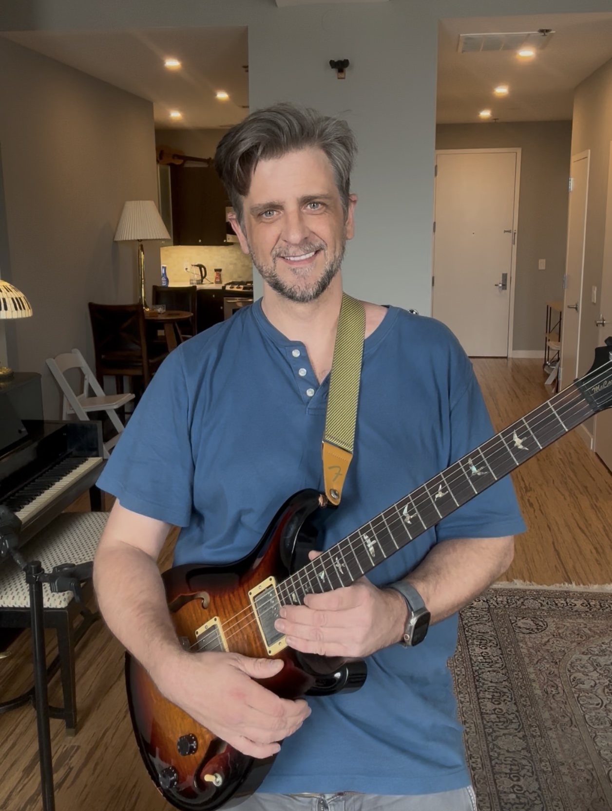 Ben smiling, holding his guitar in the studio with a grand piano behind him